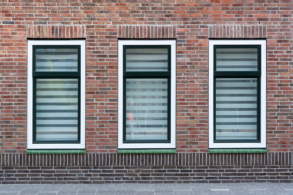 Red brick wall with three tall white-framed windows with dark inner frames and striped blinds